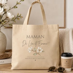 May include: A beige canvas tote bag featuring the text MAMAN De L&eacute;a et Emma with a small heart icon, a floral illustration of white magnolia blossoms, and the text MA PLUS BELLE HISTOIRE with a small heart icon centered at the bottom.