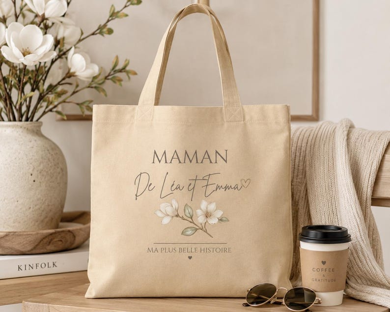 May include: A beige canvas tote bag featuring the text MAMAN De L&eacute;a et Emma with a small heart icon, a floral illustration of white magnolia blossoms, and the text MA PLUS BELLE HISTOIRE with a small heart icon centered at the bottom.