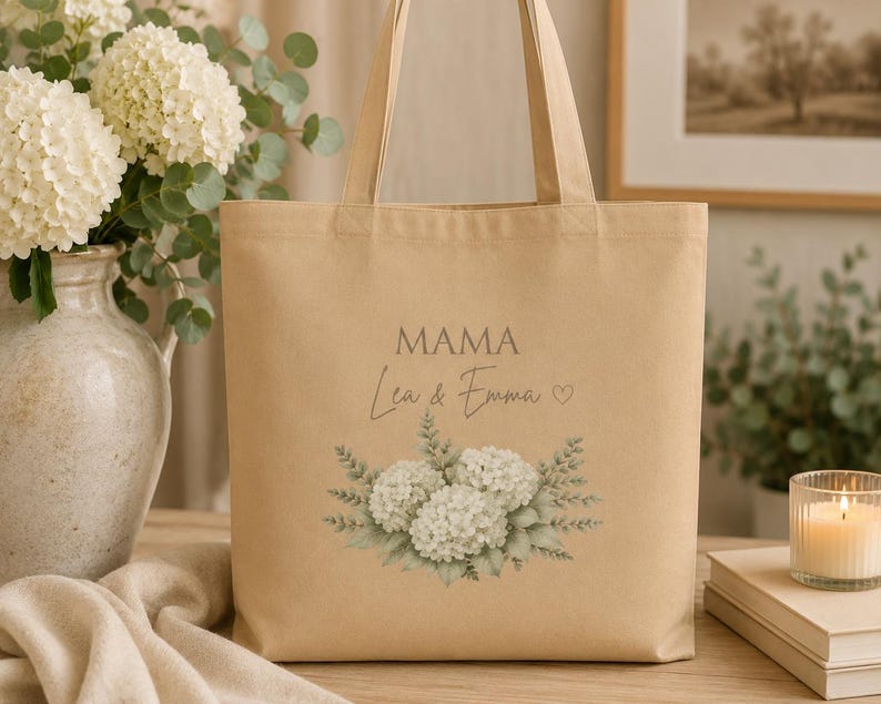 May include: A beige canvas tote bag featuring the printed text MAMA and Lea & Emma with a small heart symbol. Below the text is a detailed illustration of white hydrangea flowers surrounded by green leaves and foliage on the front panel.