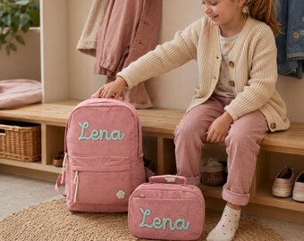 May include: A pink corduroy backpack and a matching rectangular lunch bag, both featuring the name Lena embroidered in light blue script. Each item is accented with a small white flower patch near the bottom, white zippers, and pulls.