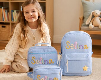 May include: A light blue corduroy backpack, a matching lunch bag, and a pencil case, each featuring the name Selina in colorful, raised, cursive lettering. The backpack includes a small yellow star patch on the front pocket.