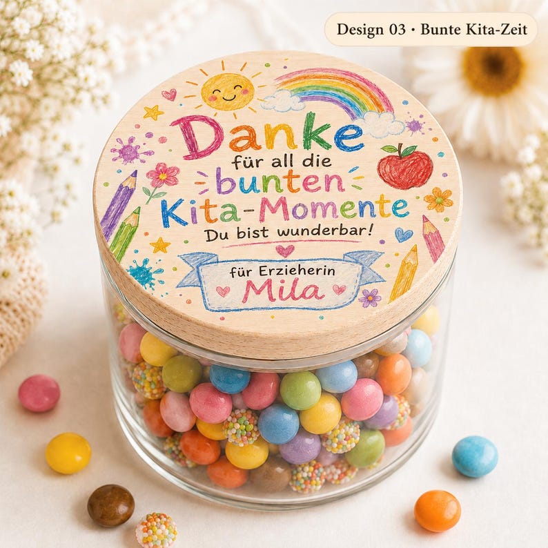 May include: A clear glass jar filled with colorful round candies, topped with a round wooden lid. The lid features a printed design with a rainbow, a sun, a red apple, pencils, and the German text: Danke f&uuml;r all die bunten Kita-Momente.