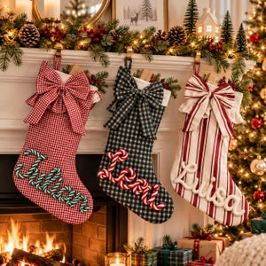 Puede incluir: Tres calcetines de Navidad colgados de una repisa. El primero es de cuadros rojos y blancos con un lazo grande y el nombre Julian en letras de bastón de caramelo verde. El segundo es de cuadros negros y blancos con un lazo y el nombre Levi en letras de bastón de caramelo rojo. El tercero es de rayas rojas y blancas con un lazo y el nombre Luisa en letras blancas.