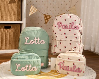 May include: Two corduroy backpacks and two matching lunch bags. One set is solid sage green with the name Lotta in pink script. The other set is cream with a red strawberry pattern and the name Paula in pink script. Each bag has a front zipper.