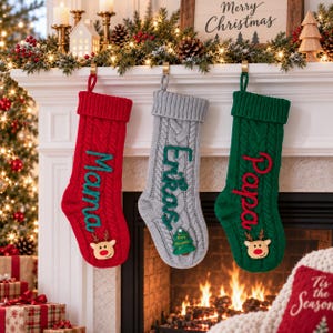 May include: Three knit Christmas stockings hanging in a row. The left is red with the name Mama and a reindeer face. The center is gray with the name Erika and a small tree. The right is green with the name Papa and a reindeer face.