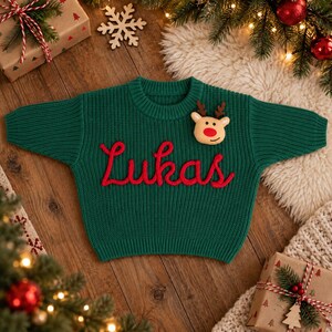 May include: A dark green long-sleeve knit sweater featuring the name Lukas embroidered in red cursive script across the chest. A small, plush reindeer head with brown antlers and a bright red nose is attached to the upper right side of the sweater.
