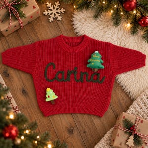 May include: A bright red knit sweater featuring the name Carina embroidered in dark green yarn across the chest. The sweater is decorated with two small, plush Christmas tree ornaments, one of which has the text Merry Christmas stitched on it.