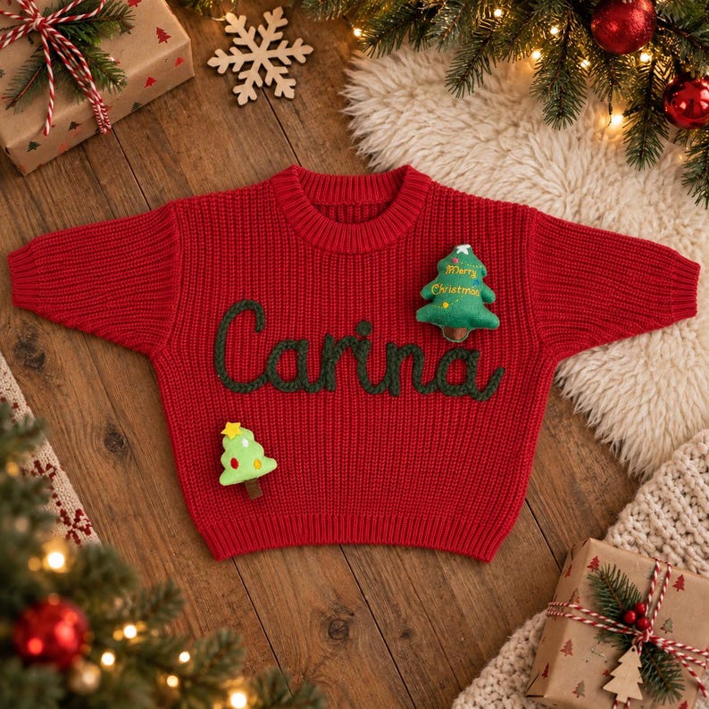 May include: A bright red knit sweater featuring the name Carina embroidered in dark green yarn across the chest. The sweater is decorated with two small, plush Christmas tree ornaments, one of which has the text Merry Christmas stitched on it.