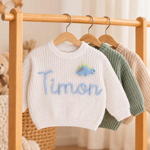 May include: A white long-sleeve knit sweater featuring the name Timon embroidered in blue script and a small blue dinosaur appliqu&eacute;. Behind it hang two additional ribbed knit sweaters in solid sage green and beige, all displayed on wooden hangers.