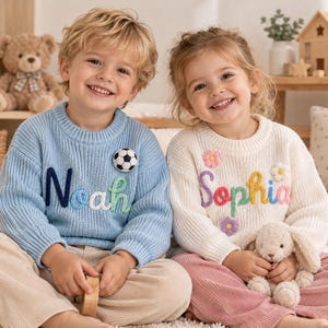May include: A light blue knit sweater featuring the name Noah in dark blue, white, and green yarn with a soccer ball patch, and a cream knit sweater featuring the name Sophia in purple, red, yellow, and pink yarn with three small flower patches.