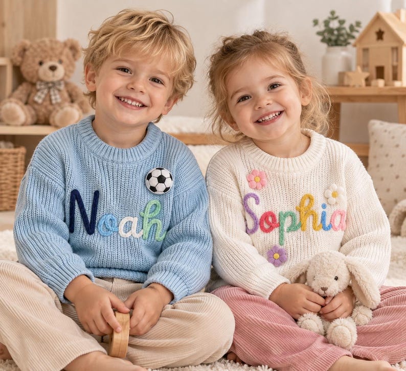 May include: A light blue knit sweater featuring the name Noah in dark blue, white, and green yarn with a soccer ball patch, and a cream knit sweater featuring the name Sophia in purple, red, yellow, and pink yarn with three small flower patches.