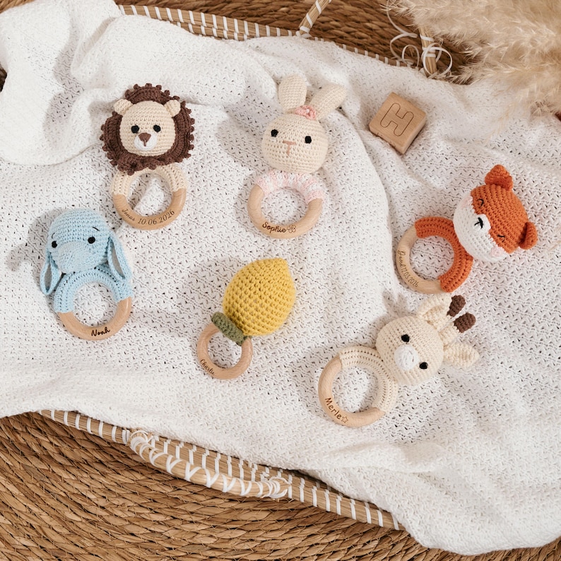 Op de afbeelding: Zes gehaakte rammelaars voor baby's in de vorm van een leeuw, konijn, olifant, citroen, vos en giraf, elk bevestigd aan een houten ring. De houten ringen zijn gegraveerd met namen en datums, waaronder Jonas, Sophie, Noah, Charlie en Merle.