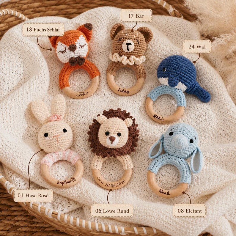 Op de afbeelding: Zes gehaakte dierenrammelaars met houten ringen. De set bevat een vos, beer, walvis, konijn, leeuw en olifant. Elke houten ring is gegraveerd met een naam of datum. De labels 18 Fuchs Schlaf, 17 B&auml;r, 24 Wal, 01 Hase Ros&eacute;, 06 L&ouml;we Rund en 08 Elefant zijn zichtbaar.