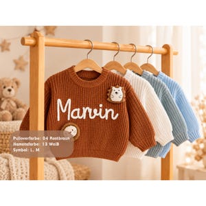 May include: A rust-brown knitted sweater featuring the white embroidered name Marvin, a hedgehog patch, and a lion patch. Behind it hang three additional sweaters in white, light blue, and a slightly darker blue on wooden hangers.