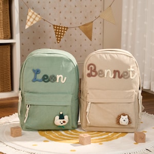 Könnte beinhalten: Zwei Rucksäcke aus Cord, einer in Salbeigrün und einer in Beige, jeweils mit einer Vordertasche und weißen Reißverschlussanhängern. Der grüne Rucksack trägt den Namen Leon in Blau und Grün, der beige Rucksack Bennet in Braun und Creme.