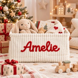 May include: A rectangular cream-colored corduroy storage basket featuring the name Amelie embroidered in red script on the front. The basket contains a plush tan bunny, a book with a Christmas tree illustration, and a white knit blanket with red hearts.