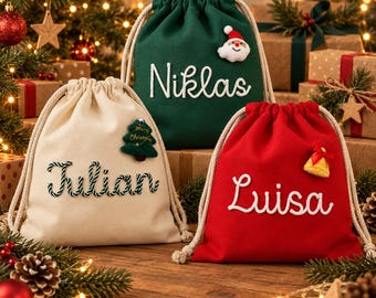 Op de afbeelding: Drie cadeauzakjes met trekkoord in crème, donkergroen en rood. Het crèmekleurige zakje heeft de naam Julian en een kerstboom-patch. Het groene zakje toont de naam Niklas en een kerstman-patch. Het rode zakje toont de naam Luisa en een bel-patch.