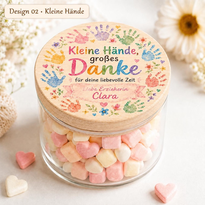 May include: A clear glass jar filled with pink, white, and peach heart-shaped candies, topped with a round wooden lid. The lid features colorful handprint illustrations, floral accents, and the German text "Kleine H&auml;nde, gro&szlig;es Danke, f&uuml;r deine liebevolle Zeit, Liebe Erzieherin Clara."