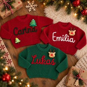 May include: Three knitted sweaters in red and green with long sleeves and crew necks. Two red sweaters feature the names Carina in green and Emilia in white, while the green sweater features the name Lukas in red, with festive appliques.