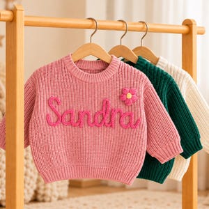 May include: A pink knit sweater featuring the name Sandra embroidered in cursive yarn lettering and a small pink flower appliqué on the chest, hanging alongside a dark green knit sweater and a cream knit sweater on a wooden clothing rack.