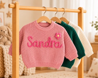 May include: A pink knit sweater featuring the name Sandra embroidered in cursive yarn lettering and a small pink flower appliqué on the chest, hanging alongside a dark green knit sweater and a cream knit sweater on a wooden clothing rack.