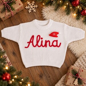 May include: A white long-sleeve knit sweater featuring the name Alina embroidered in red cursive script across the chest. A small red felt Santa hat applique with a white snowflake detail is positioned above the letter i in the name.