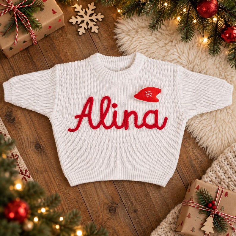 May include: A white long-sleeve knit sweater featuring the name Alina embroidered in red cursive script across the chest. A small red felt Santa hat applique with a white snowflake detail is positioned above the letter i in the name.