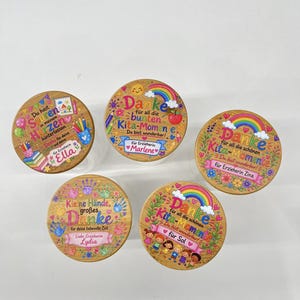 Puede incluir: Cinco tapas redondas de madera para frascos con dise&ntilde;os impresos coloridos, texto en alem&aacute;n, arco&iacute;ris, flores y &uacute;tiles escolares. Cada tapa est&aacute; personalizada con un nombre, incluyendo Ella, Marlene, Zina, Lydia y Sol, para docentes.