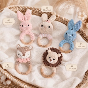 Op de afbeelding: Vijf gehaakte rammelaars met houten ringen, waaronder een roze konijn gelabeld Hase Rosa, een beige konijn gelabeld Hase Ros&eacute;, een blauw konijn gelabeld Hase Blau, een ree gelabeld Reh Ros&eacute; en een leeuw gelabeld L&ouml;we Rund, met namen in de ringen gegraveerd.