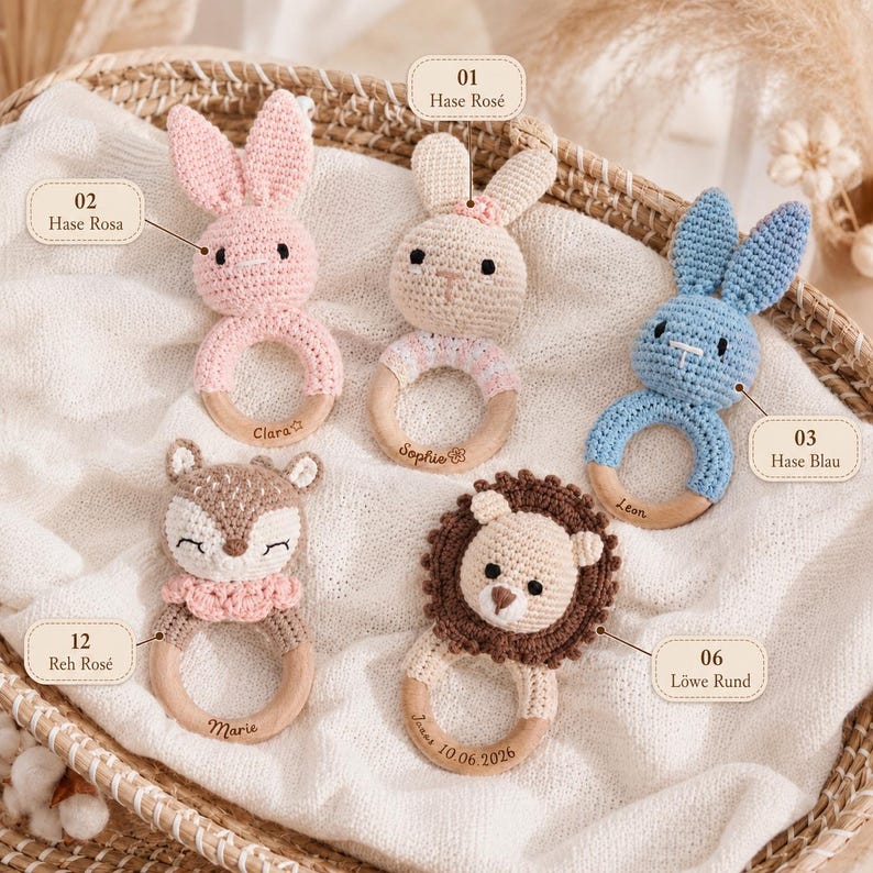 Op de afbeelding: Vijf gehaakte rammelaars met houten ringen, waaronder een roze konijn gelabeld Hase Rosa, een beige konijn gelabeld Hase Ros&eacute;, een blauw konijn gelabeld Hase Blau, een ree gelabeld Reh Ros&eacute; en een leeuw gelabeld L&ouml;we Rund, met namen in de ringen gegraveerd.