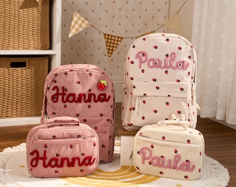 May include: Two corduroy backpacks and two matching lunch bags featuring a strawberry pattern. One set is pink with the name Hanna embroidered in red, and the other is cream with the name Parila embroidered in pink. Each bag has a zippered front pocket.