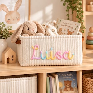 Puede incluir: Una cesta de almacenamiento de pana color crema con el nombre Luisa bordado en hilo de colores pastel. La cesta contiene un conejo de peluche beige, una manta con estampado floral y un libro titulado Meine liebsten Tierfreunde.