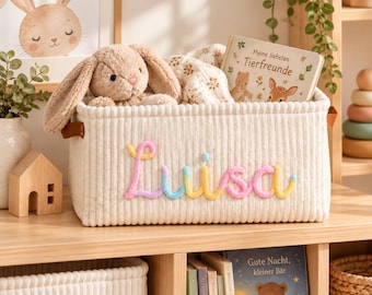 Op de afbeelding: Een crèmekleurige opbergmand van corduroy met de naam Luisa geborduurd in pasteltinten. De mand bevat een beige pluche konijn, een deken met bloemenpatroon en een boek met de titel Meine liebsten Tierfreunde.
