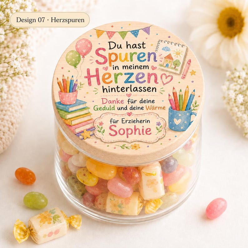 May include: A clear glass jar filled with colorful jelly beans, topped with a round wooden lid. The lid features a printed design with the German text 'Du hast Spuren in meinem Herzen hinterlassen', illustrations of balloons, books, pencils, and a mug.