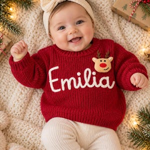 May include: A red long-sleeved knit sweater featuring the name Emilia embroidered in white script across the chest, accented with a small plush reindeer applique with a red nose. The sweater is paired with cream-colored ribbed leggings.