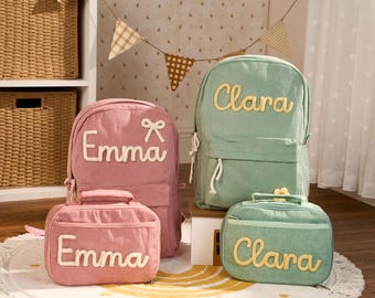May include: Two corduroy backpacks and two matching lunch bags in dusty rose and sage green. The rose items are personalized with the name Emma in white, and the green items feature the name Clara in yellow. Each bag has a front zipper pocket.