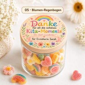 Puede incluir: Un frasco de vidrio transparente lleno de caramelos de goma coloridos con formas de corazones, flores y arco&iacute;ris. La tapa de madera presenta un arco&iacute;ris impreso, detalles florales y el texto Danke f&uuml;r all die sch&ouml;nen Kita-Momente.