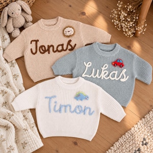 May include: Three long-sleeve knit sweaters in beige, light blue, and white. The beige sweater features the name Jonas and a lion patch. The light blue sweater displays the name Lukas and a red car patch. The white sweater shows the name Timon and a blue dinosaur patch.