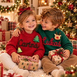May include: A red knitted sweater with the name Carina embroidered in green and a green knitted sweater with the name Lukas embroidered in red. Both sweaters feature festive felt appliques of a Christmas tree and a reindeer respectively.
