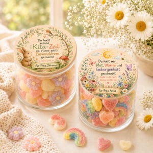 May include: Two clear glass jars filled with colorful, sugar-coated gummy candies in shapes of flowers and rainbows. Each jar has a wooden lid featuring printed illustrations of nature, a rabbit, and German text, personalized for Frau Johanna and Frau Anna.