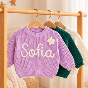 May include: A lavender long-sleeve knit sweater featuring the name Sofia in white cursive lettering and a small white flower appliqu&eacute; on the chest. The sweater hangs on a wooden hanger alongside a dark green sweater and a cream-colored sweater.