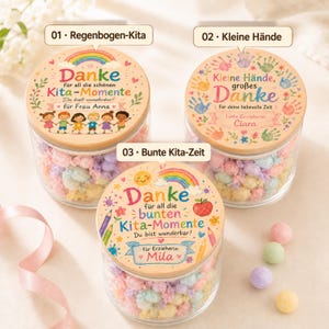Puede incluir: Tres frascos de vidrio transparente con tapas de madera, llenos de dulces envueltos en colores pastel. Las tapas presentan ilustraciones coloridas de arco&iacute;ris, ni&ntilde;os, huellas de manos y manzanas, con texto en alem&aacute;n que incluye 'Danke' y 'Kita-Momente'.