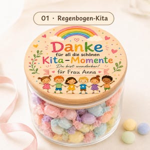 Puede incluir: Un frasco de vidrio transparente lleno de dulces envueltos en colores pastel, con una tapa redonda de madera. La tapa presenta un arco&iacute;ris colorido, seis ni&ntilde;os ilustrados tomados de la mano y el texto en alem&aacute;n Danke f&uuml;r all die sch&ouml;nen Kita-Momente, Du bist wunderbar, f&uuml;r Frau Anna.