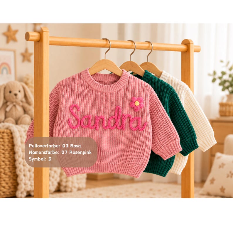 May include: A pink knit sweater featuring the name Sandra in raised pink yarn lettering and a small pink flower appliqu&eacute; with a yellow center. The sweater is displayed on a wooden hanger alongside a dark green sweater and a cream-colored sweater.