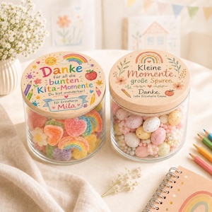 May include: Two clear glass jars with round wooden lids. The left lid features a rainbow, sun, apple, and text reading Danke f&uuml;r all die bunten Kita-Momente Du bist wunderbar! f&uuml;r Erzieherin Mila. The right lid shows a rainbow, apple, and text reading Kleine Momente, gro&szlig;e Spuren. Danke, liebe Erzieherin Emma. The jars are filled with colorful gummy candies and speckled chocolate eggs.