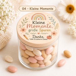 Puede incluir: Un frasco de vidrio transparente lleno de dulces ovalados en tonos rosa, crema y beige. La tapa de madera presenta un arco&iacute;ris, flores, una manzana, un l&aacute;piz y el texto en alem&aacute;n: Kleine Momente, gro&szlig;e Spuren. Danke, liebe Erzieherin Emma.