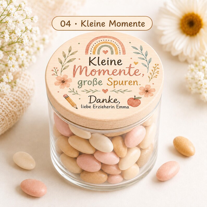 Puede incluir: Un frasco de vidrio transparente lleno de dulces ovalados en tonos rosa, crema y beige. La tapa de madera presenta un arco&iacute;ris, flores, una manzana, un l&aacute;piz y el texto en alem&aacute;n: Kleine Momente, gro&szlig;e Spuren. Danke, liebe Erzieherin Emma.