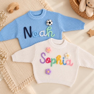 May include: A light blue knit sweater with the name Noah in dark blue, white, and green yarn, featuring a soccer ball patch. A white knit sweater with the name Sophia in purple, pink, green, yellow, and blue yarn, featuring three flower patches.