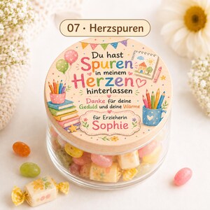 Puede incluir: Un frasco de vidrio transparente lleno de dulces coloridos, con una tapa redonda de madera. La tapa presenta un dise&ntilde;o impreso con libros, l&aacute;pices, globos y el texto en alem&aacute;n: Du hast Spuren in meinem Herzen hinterlassen.