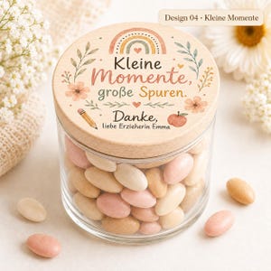 May include: A clear glass jar filled with oval-shaped candies in shades of pink, cream, and tan. The jar has a round wooden lid printed with a rainbow, floral accents, a pencil, an apple, and the German text Kleine Momente, gro&szlig;e Spuren. Danke, liebe Erzieherin Emma.